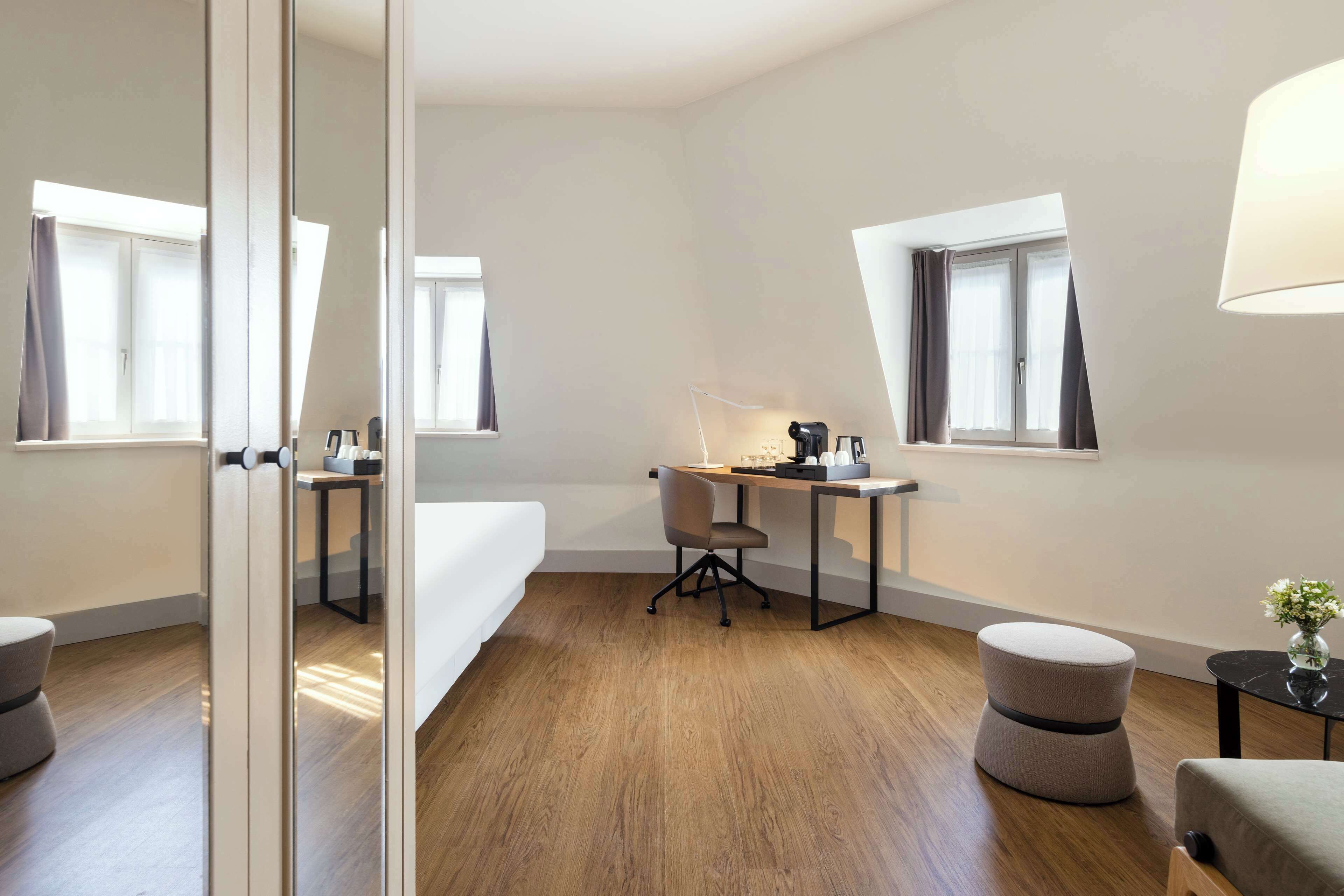 A modern hotel room with a bed, a desk, a chair, and a small table with a lamp. There are two windows with curtains, and the room has wooden flooring.