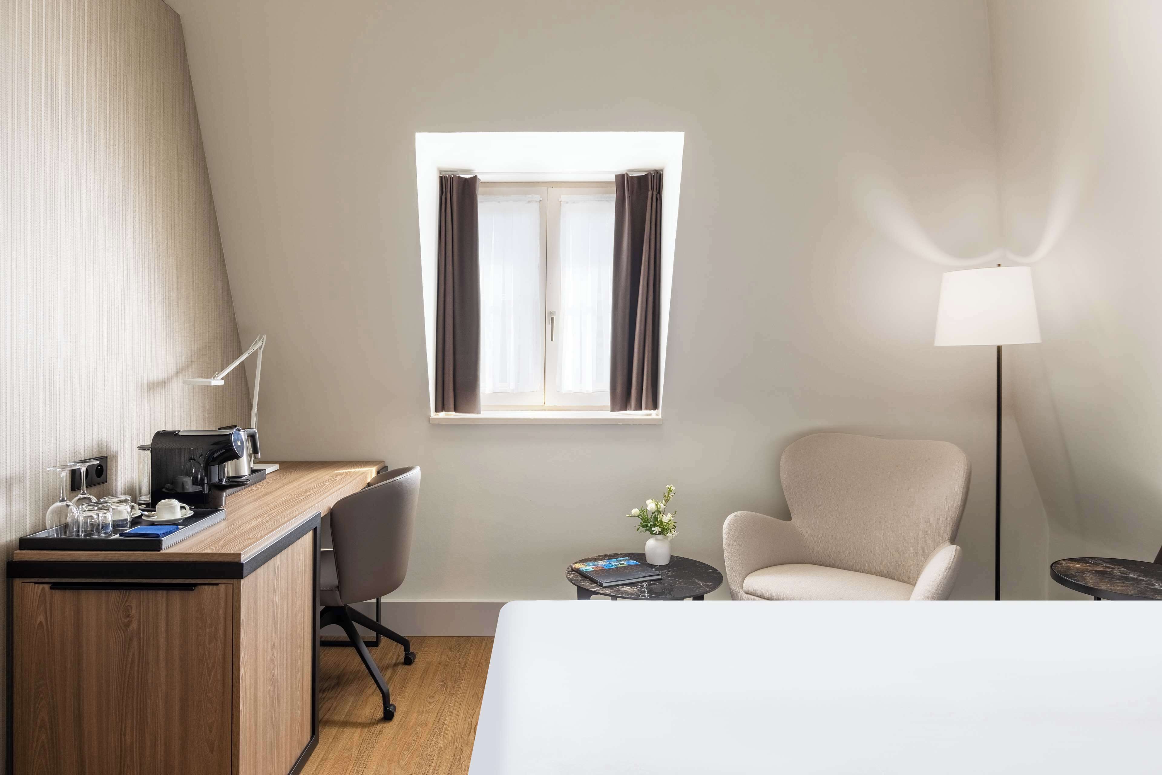 A hotel room with a bed, a desk with a coffee maker, a chair, a small table with a vase, and a lamp.