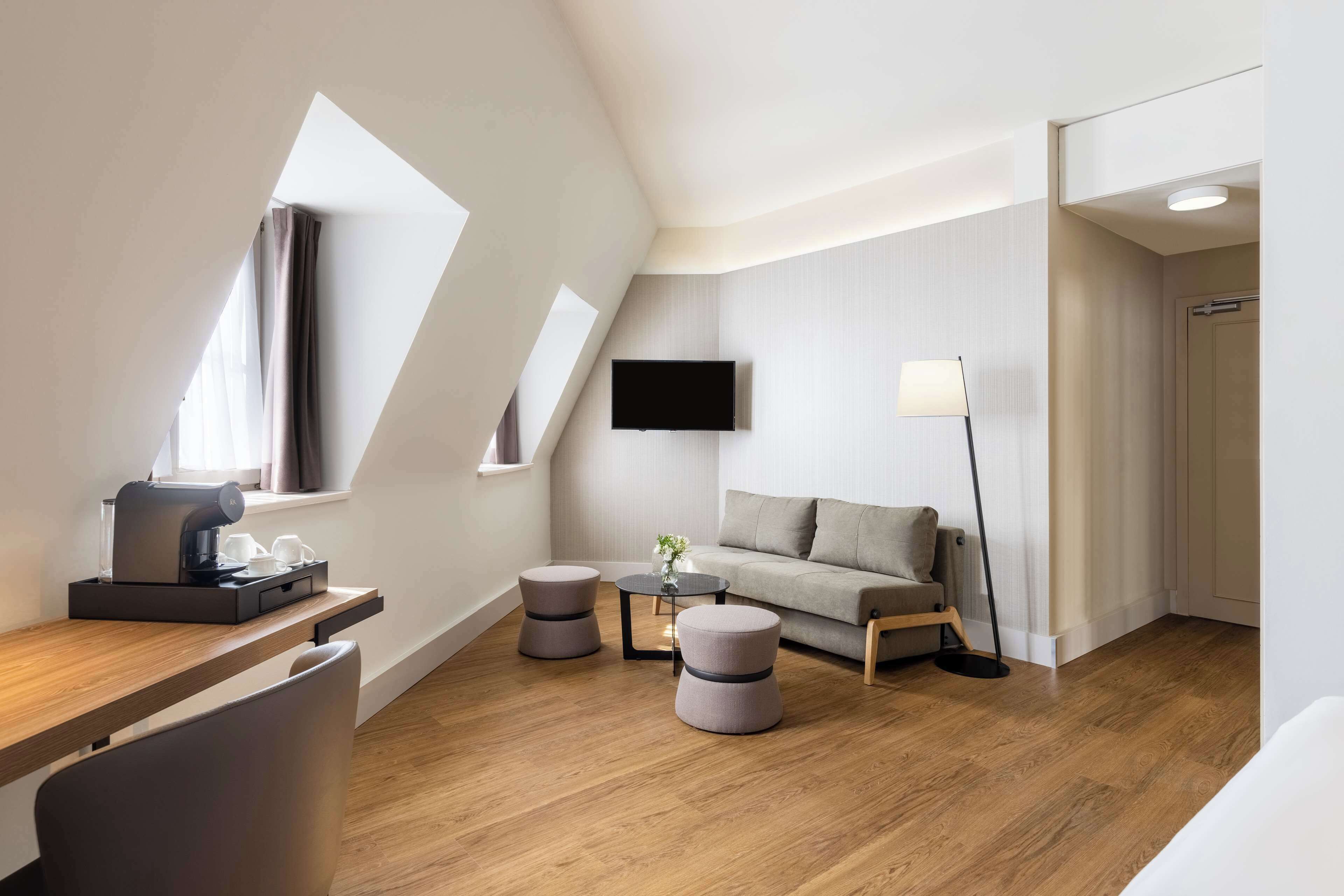 A modern hotel room with a wooden floor, a sofa, a coffee machine, a TV, and a lamp.