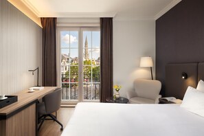 A hotel room with a bed, a desk, a chair, a lamp, and a view of a cityscape through large windows. - NH Brussels Carrefour de l'Europe (Brussels)
