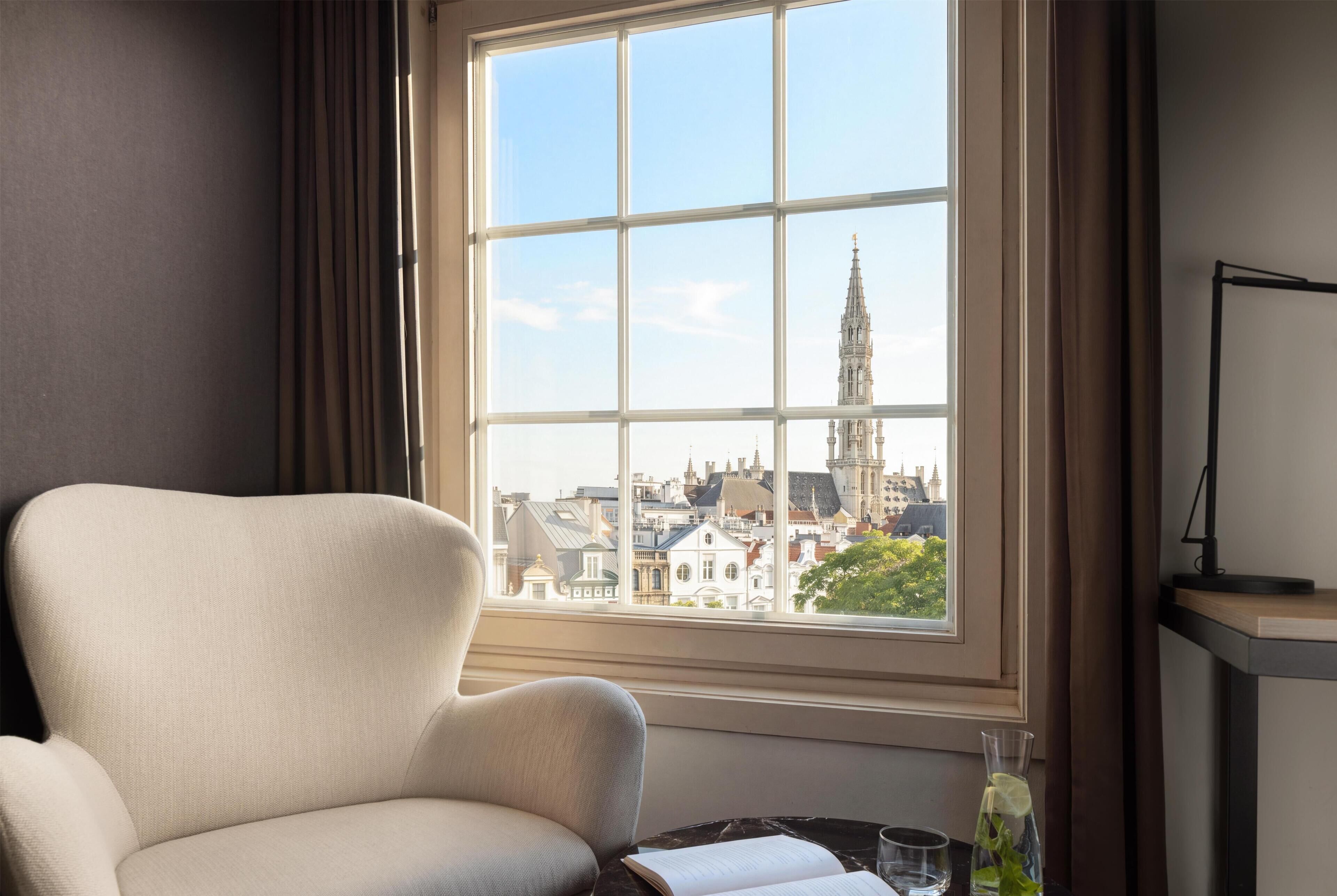 A room with a large window, a chair, a table with a book and a glass, and a lamp.