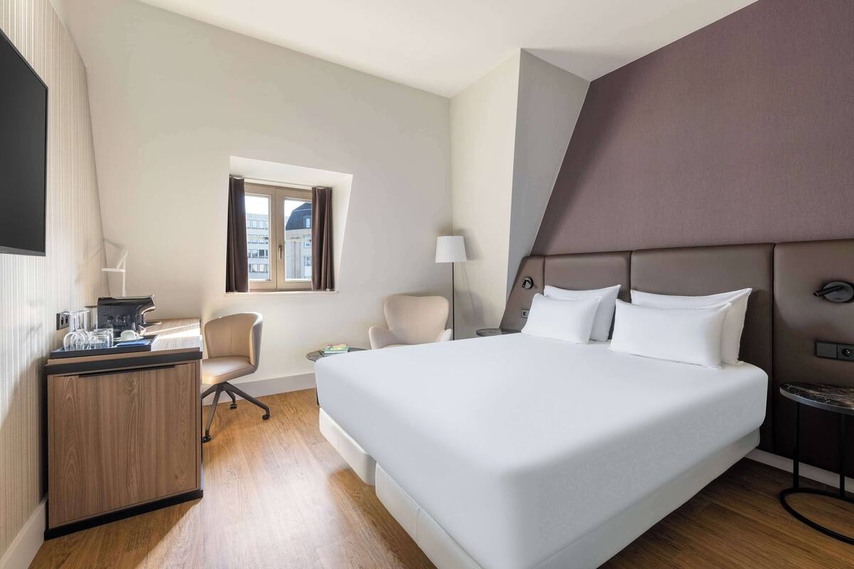 Une chambre d’hôtel moderne avec un grand lit, un bureau équipé d’un ordinateur, une chaise, une lampe et une fenêtre avec des rideaux.
