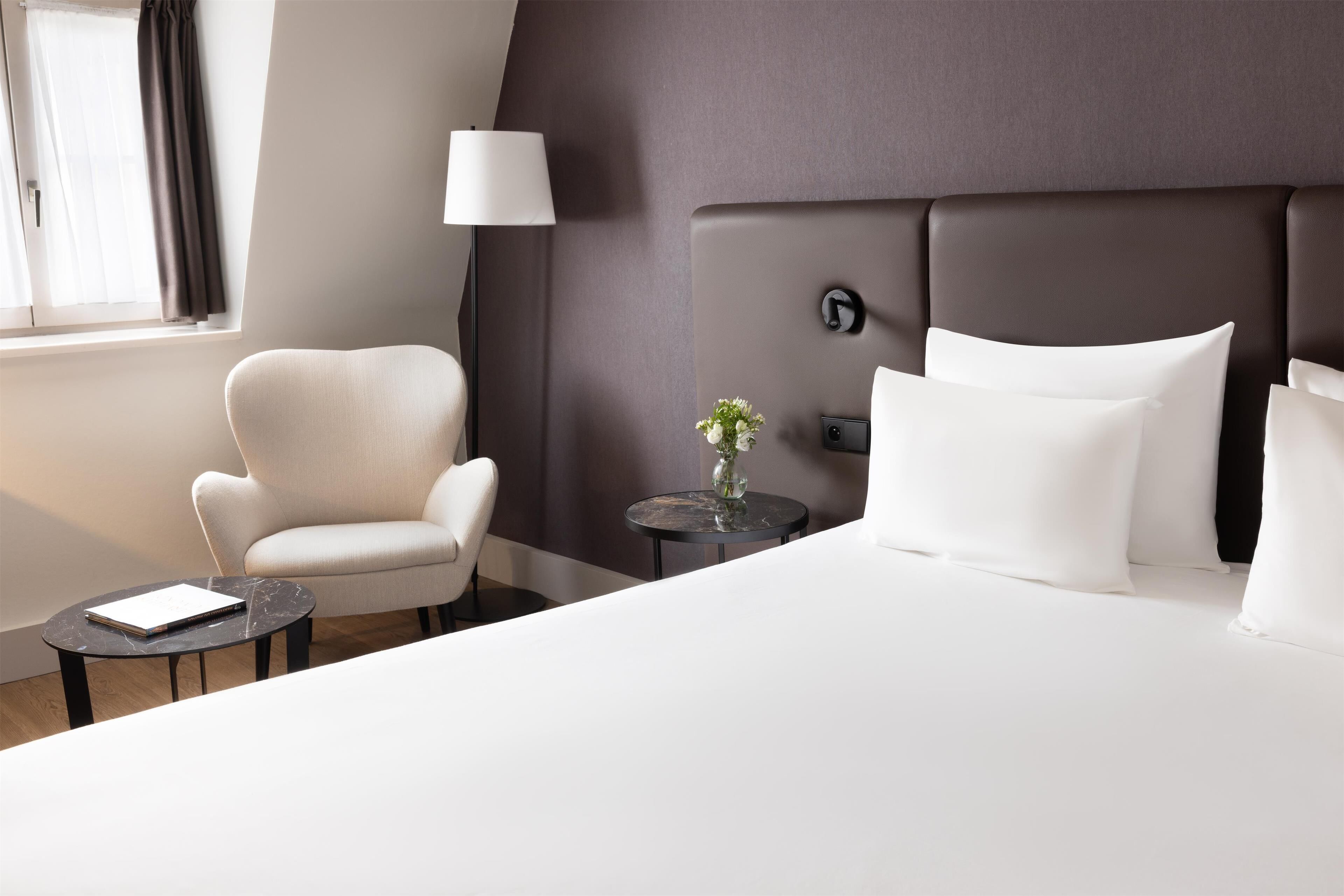 A modern hotel room with a large bed, a white armchair, a small round table with a book, a vase with flowers, and a lamp.