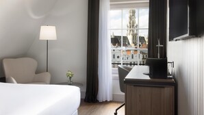 Une chambre d’hôtel comprenant un lit, un bureau, une chaise, une lampe et une grande fenêtre donnant sur un paysage urbain.