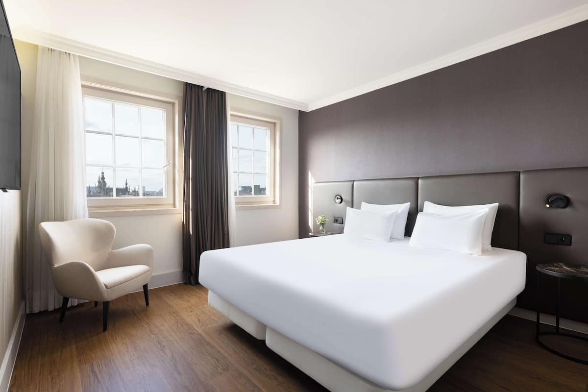 Une chambre d’hôtel moderne, dotée d’un grand lit, d’une chaise, d’une table de chevet et offrant une vue sur la ville par la fenêtre.