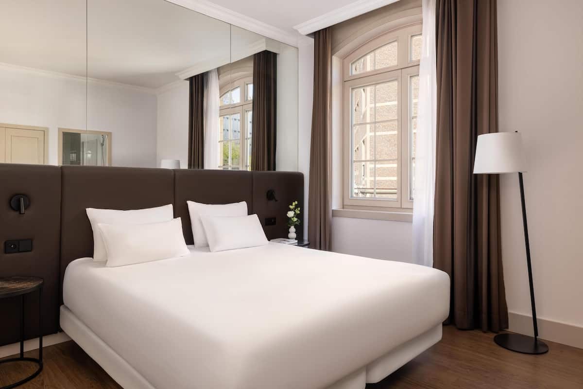 Une chambre d’hôtel moderne avec un grand lit, une table de chevet, une lampe sur pied et une fenêtre avec des rideaux.