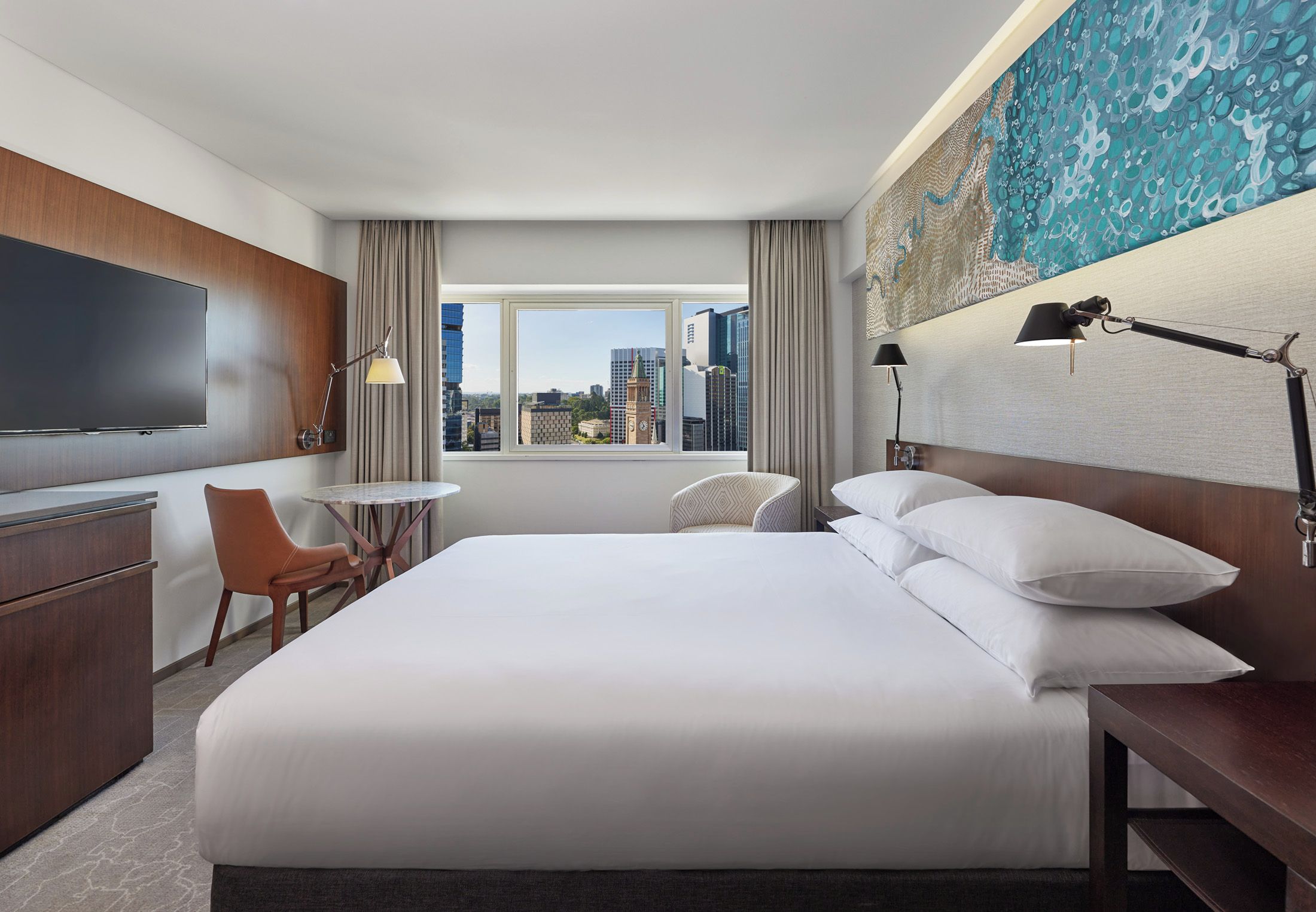A hotel room with a bed, a desk, a chair, a TV, and a view of the cityscape.