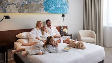 A family of four in a hotel room with a large bed, a nightstand with a lamp, a decorative wall piece, and a window with curtains.