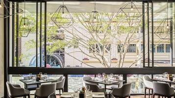 A restaurant with tables set for dining, large windows with a view of trees and buildings, and hanging geometric light fixtures.