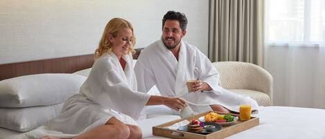 A couple in white bathrobes sitting on a bed with breakfast items including pancakes, fruit, and juice.