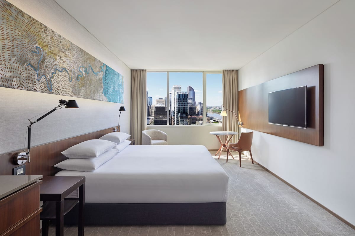A hotel room with a large bed, a desk, a chair, a TV mounted on the wall, and a window with a city view.