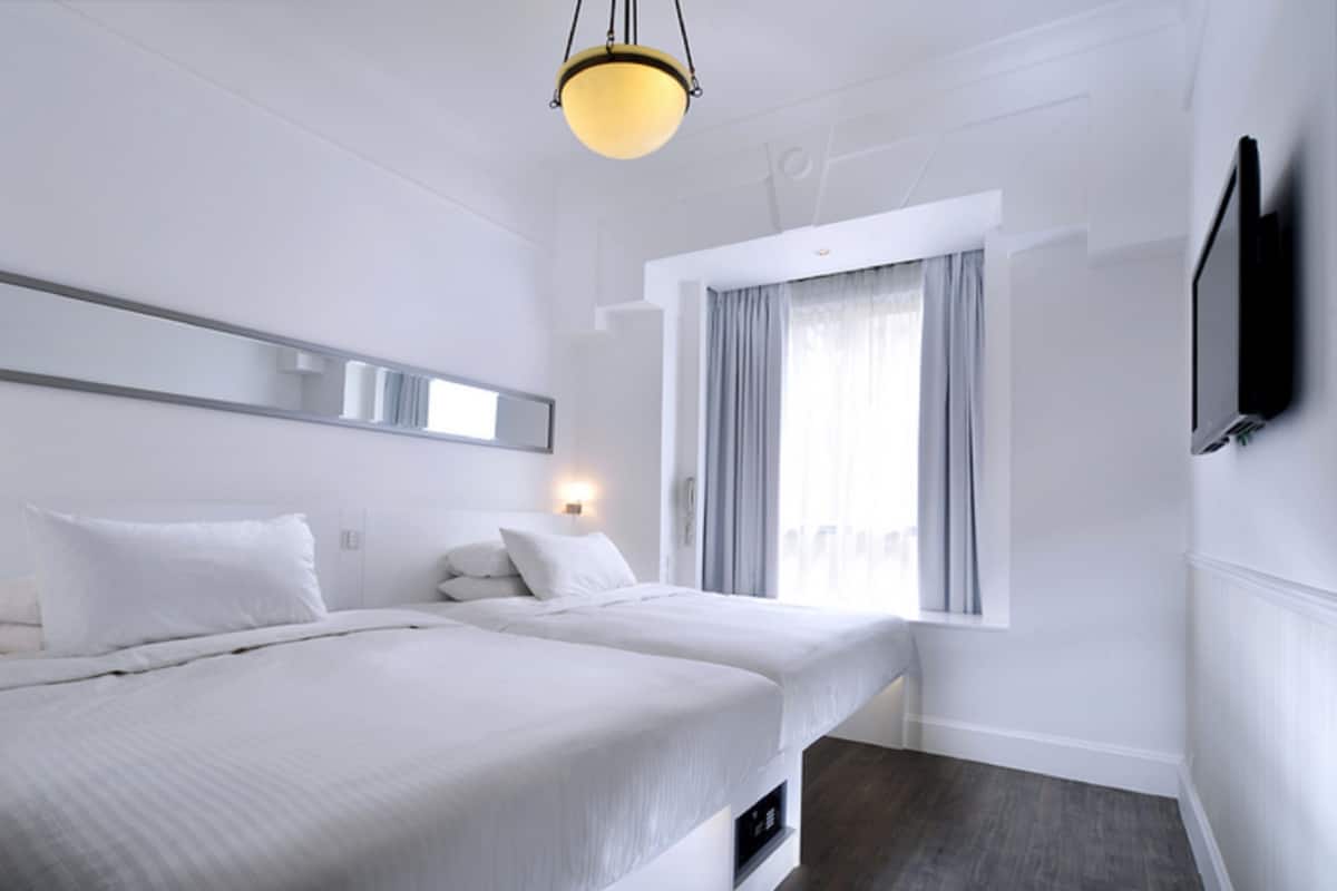 Quarto de hotel moderno com duas camas, uma janela grande com cortinas, uma TV de tela plana presa na parede e um lustre no teto.