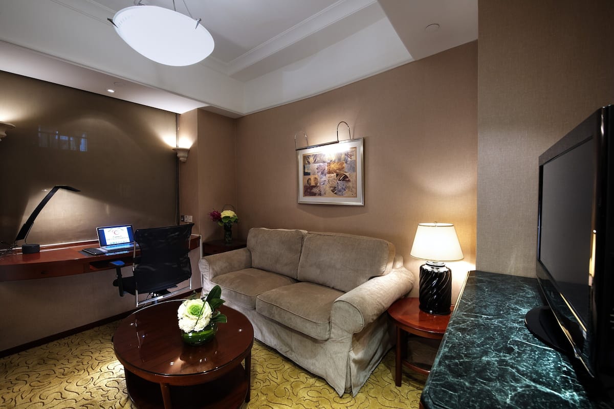 Quarto de hotel com sofá, escrivaninha com laptop, televisão e luminária.