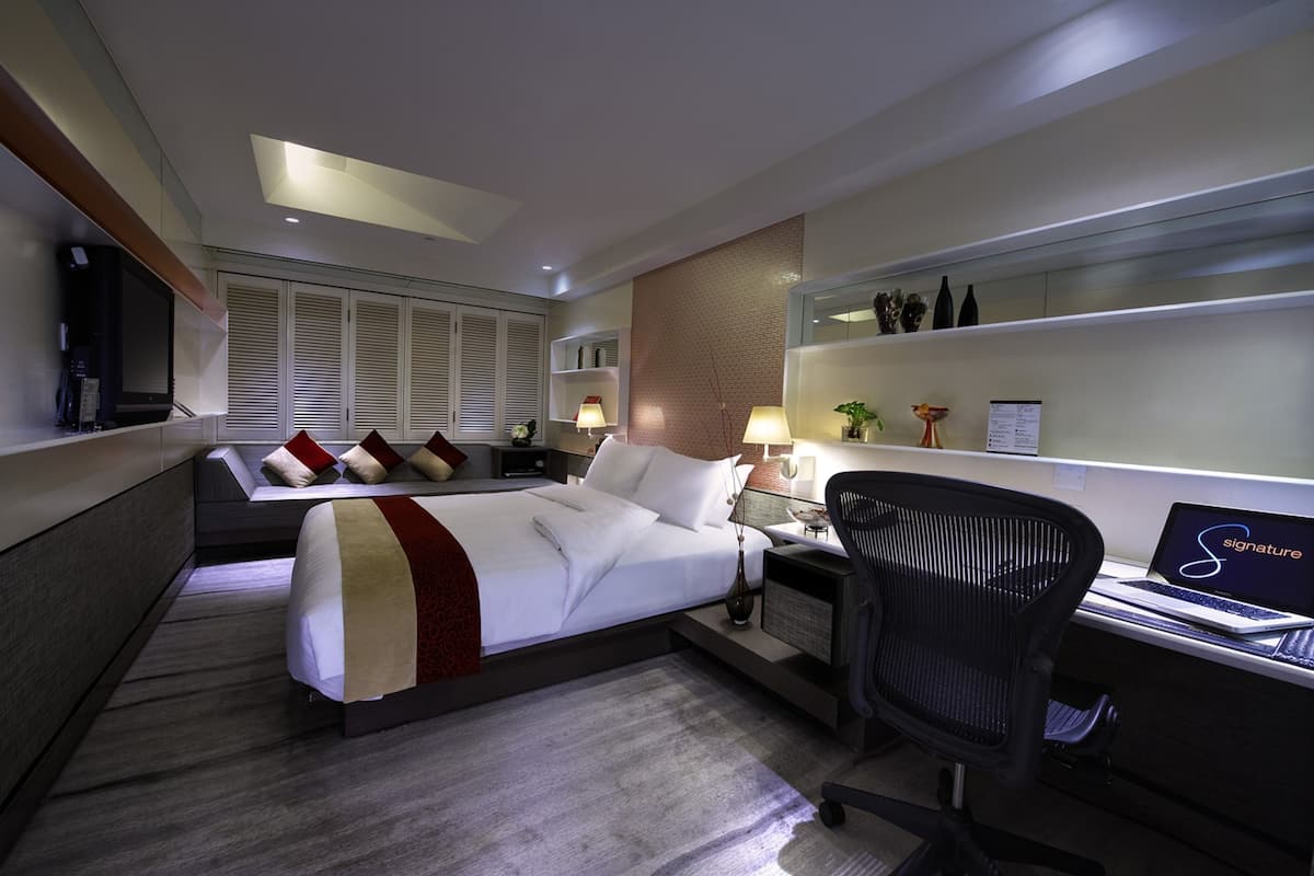 Quarto de hotel com cama, escrivaninha com laptop, cadeira, mesa de cabeceira e criado-mudo.