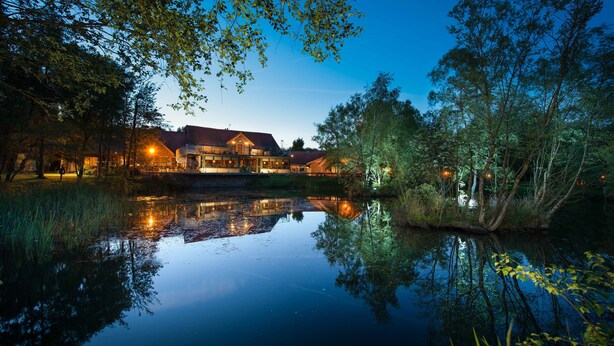 A lakeside hotel with a reflection in the water, surrounded by trees and a well-lit building.