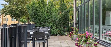 An outdoor patio with tables and chairs, a glass wall, and a hedge.