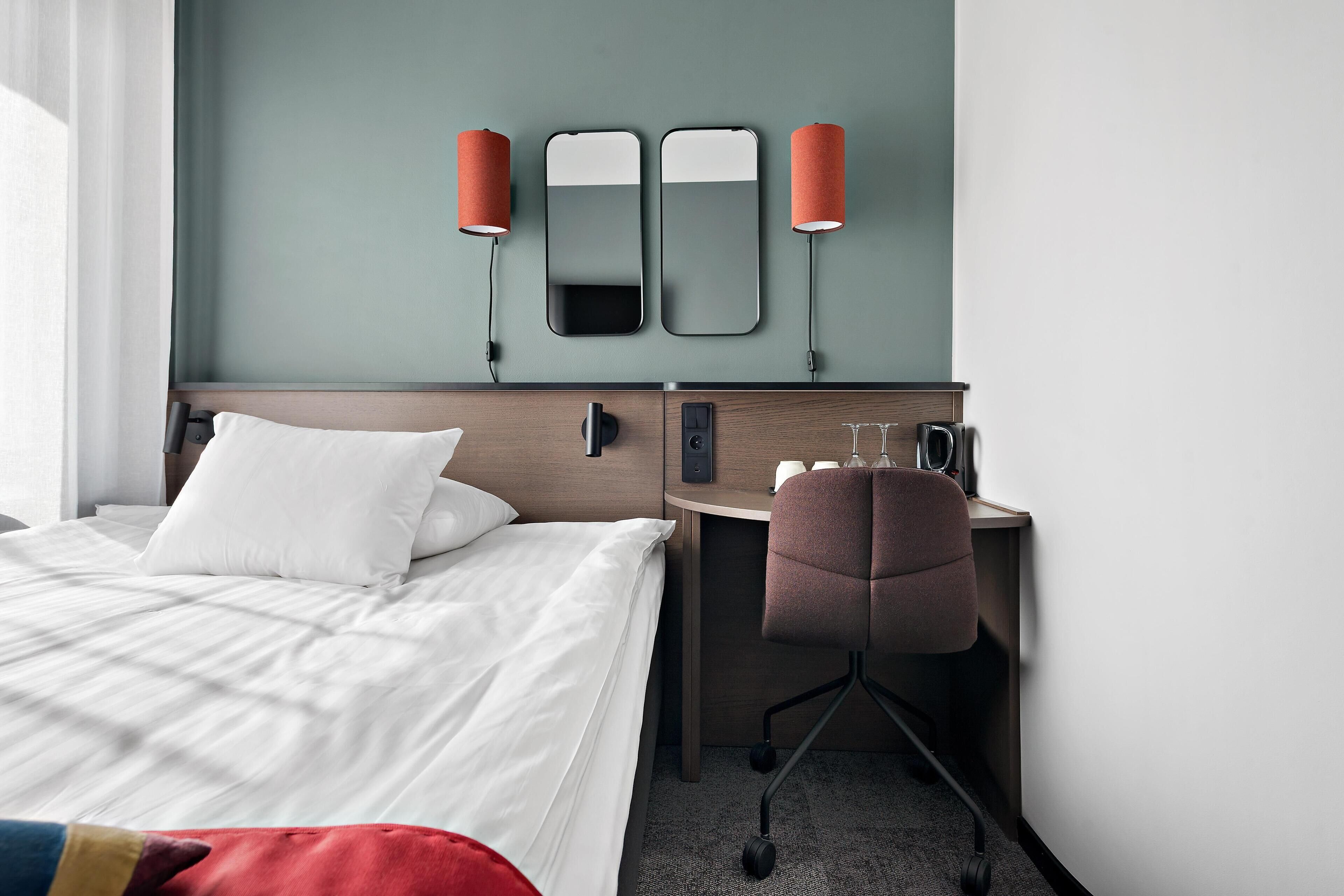 Habitación de hotel con cama, escritorio, silla, espejo y dos lámparas adosadas a la pared.