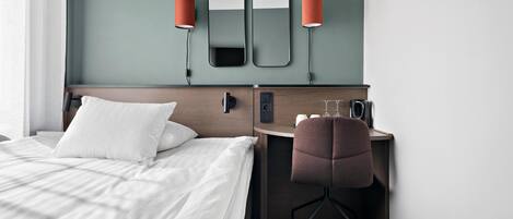 Habitación de hotel con cama, escritorio, silla, espejo y dos lámparas adosadas a la pared.