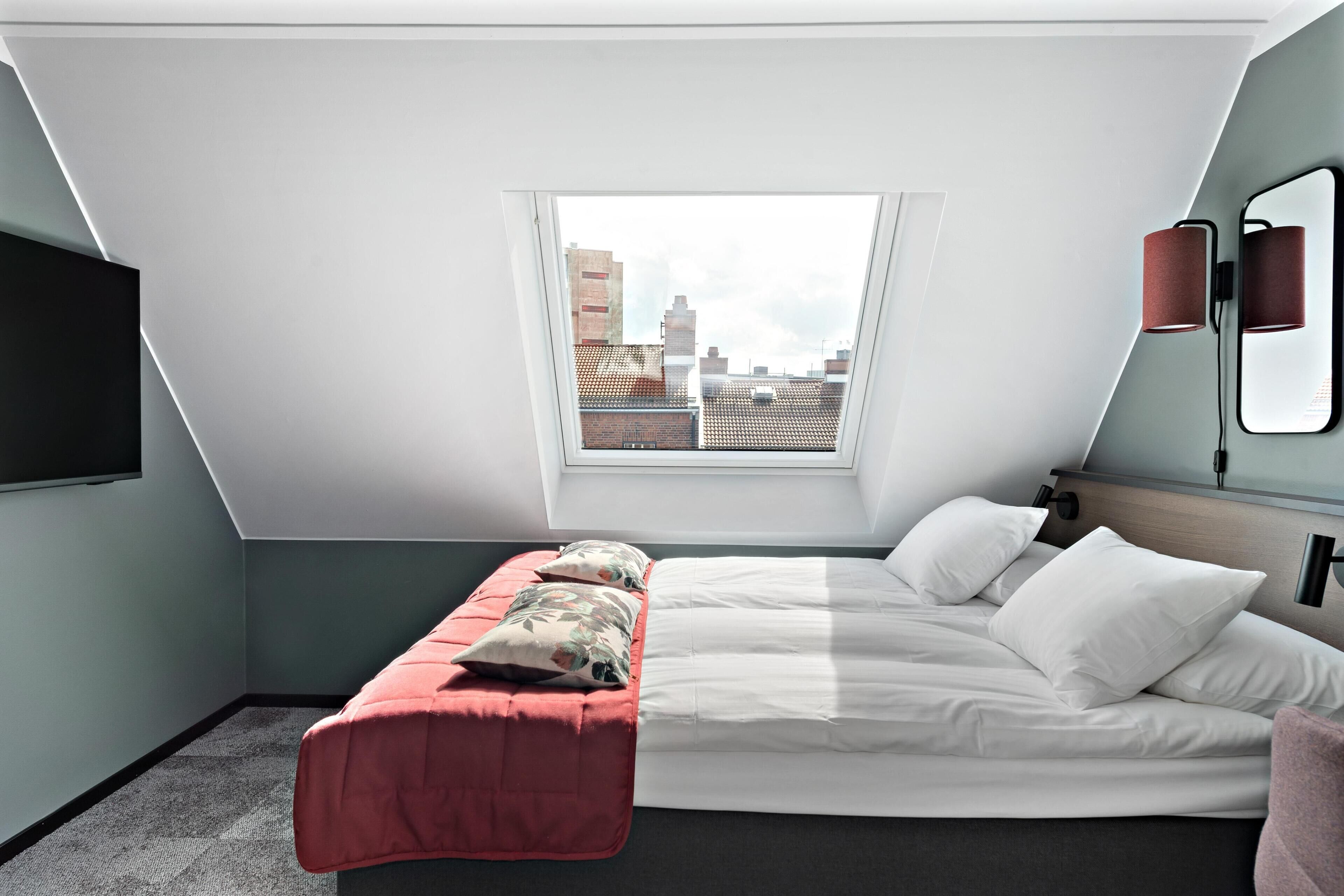A modern bedroom with a bed, a red bench, a TV, a window with a city view, and a mirror.
