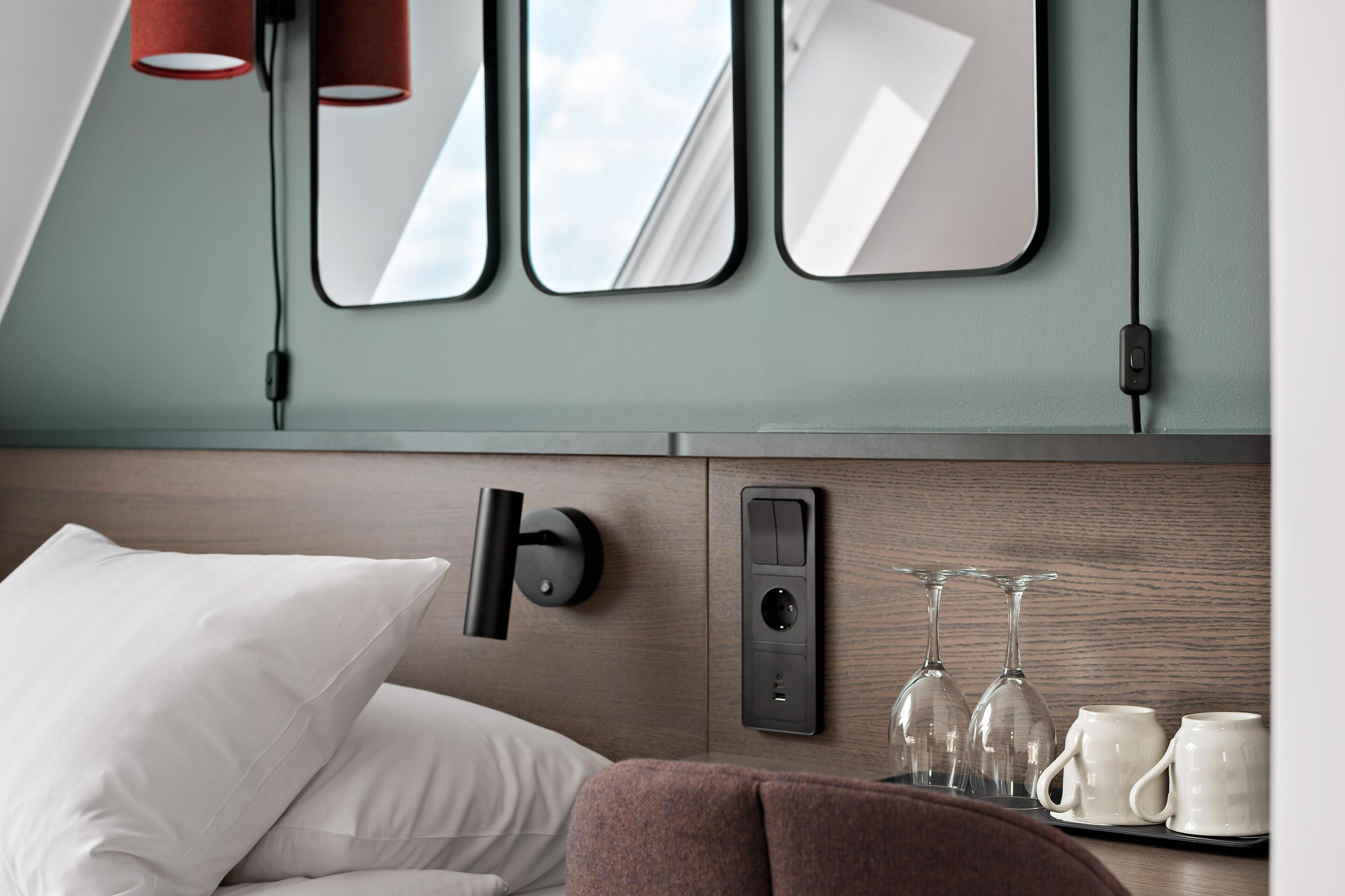 Une chambre d’hôtel moderne équipée d’un lit, d’un luminaire fixé au mur, d’une prise de courant et d’un plateau avec deux verres et deux tasses.
