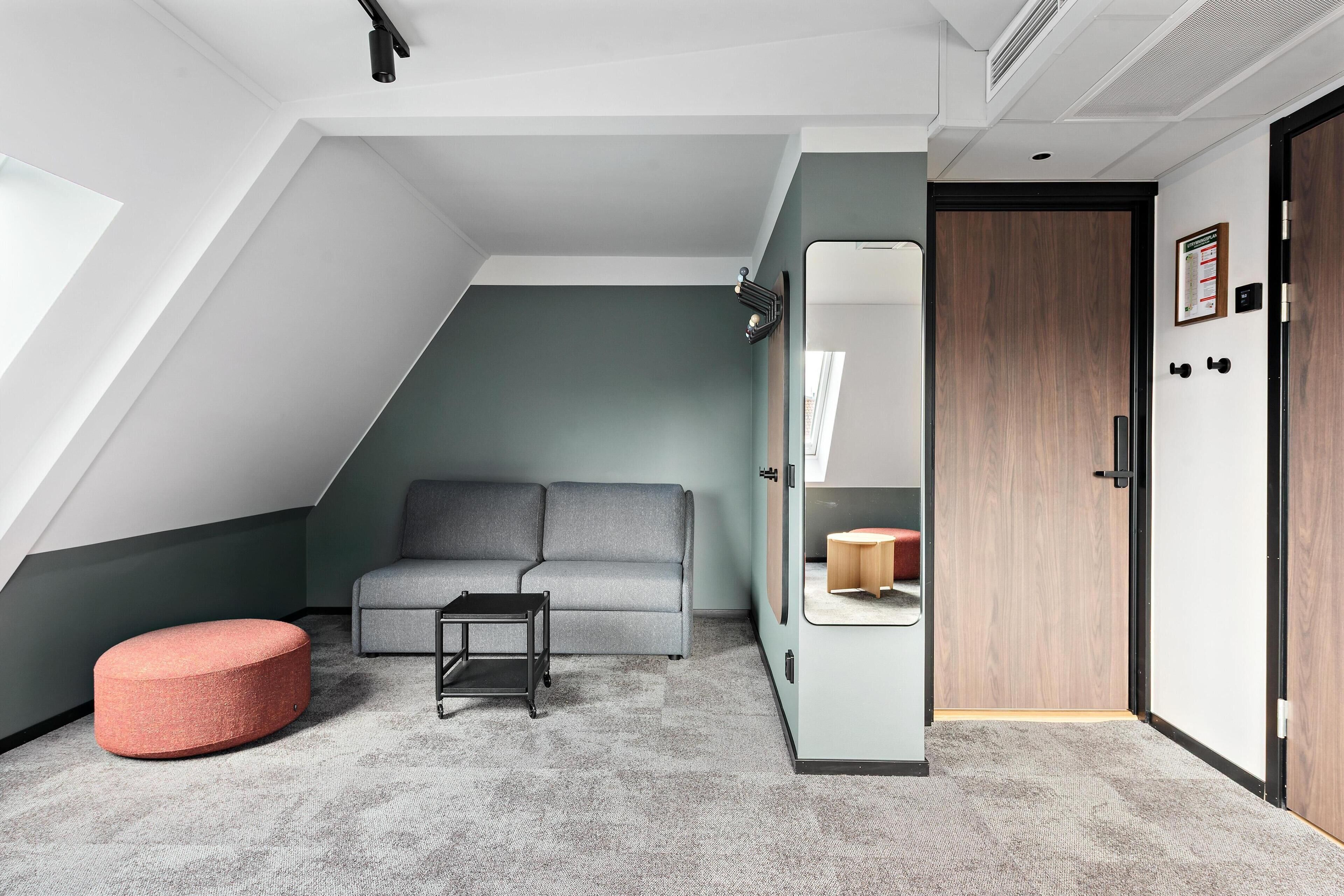 A modern room with a grey sofa, a red ottoman, a small table, a wardrobe, and a door with a mirror.