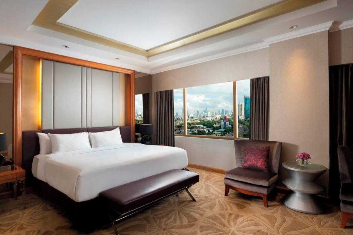 A hotel room with a large bed, a chair, a small table, and a view of a cityscape through large windows.