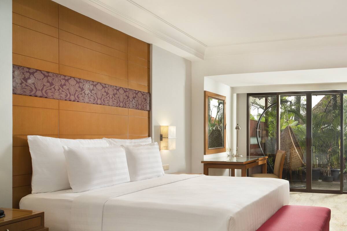 A hotel room with a large bed, a desk, a chair, and a view of the outdoors through a glass door.