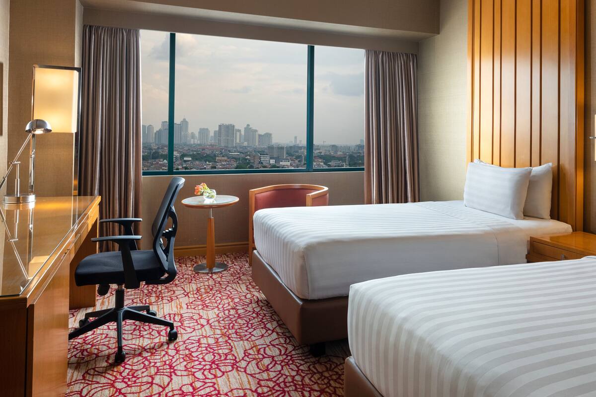 A hotel room with two beds, a desk, a chair, a small table with a vase, and a view of a cityscape through large windows.