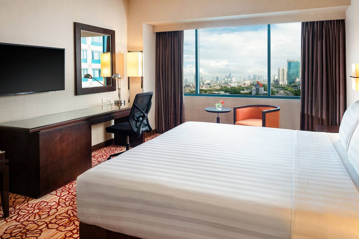 A hotel room with a large bed, a desk with a chair, a TV, and a window with a city view.