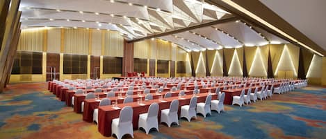 A large conference room with rows of tables and chairs, high ceilings with geometric patterns, and a carpeted floor.
