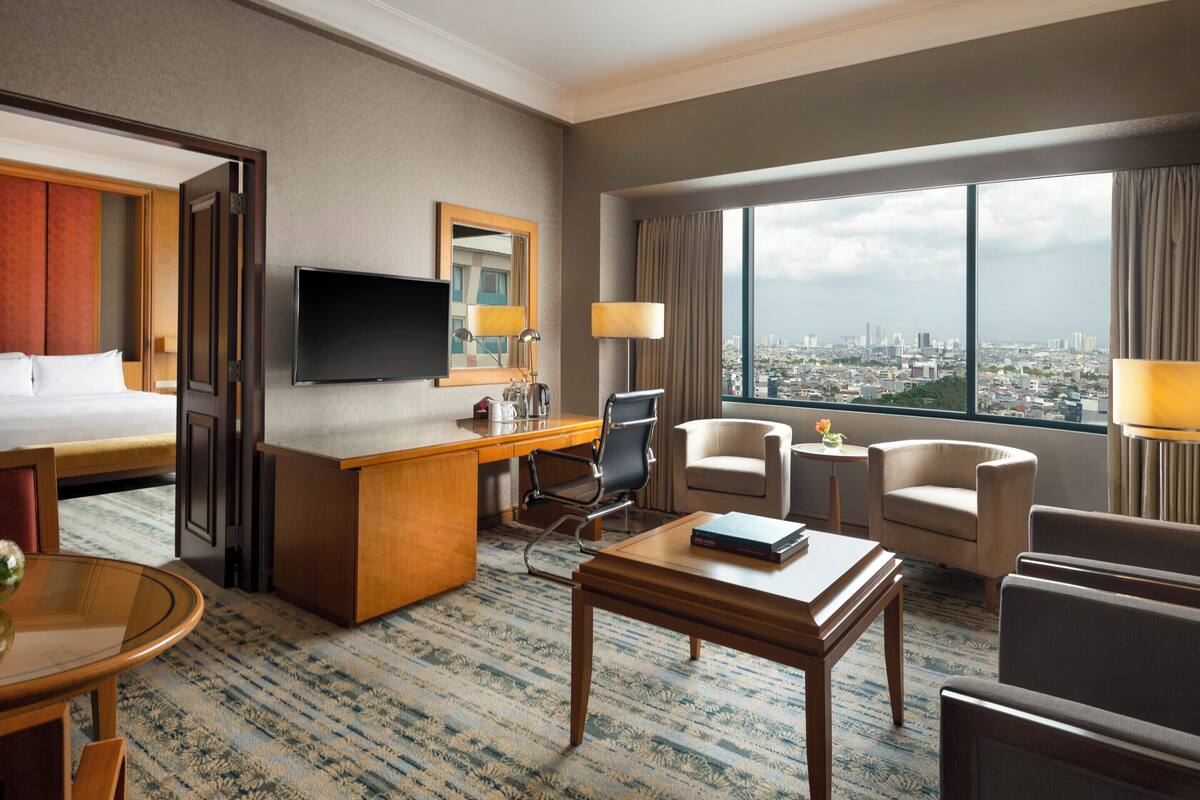 A hotel room with a bed, desk, chairs, a TV, and a view of a cityscape.