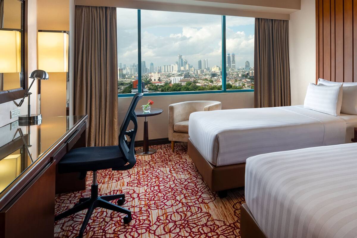 A hotel room with a city view, a desk with a lamp, a chair, a bed, and a small table with a flower vase.