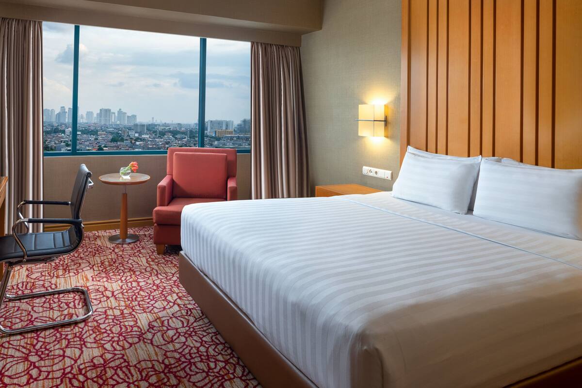 A hotel room with a large bed, a chair, a small table with a vase of flowers, a nightstand, and a view of a cityscape through large windows.