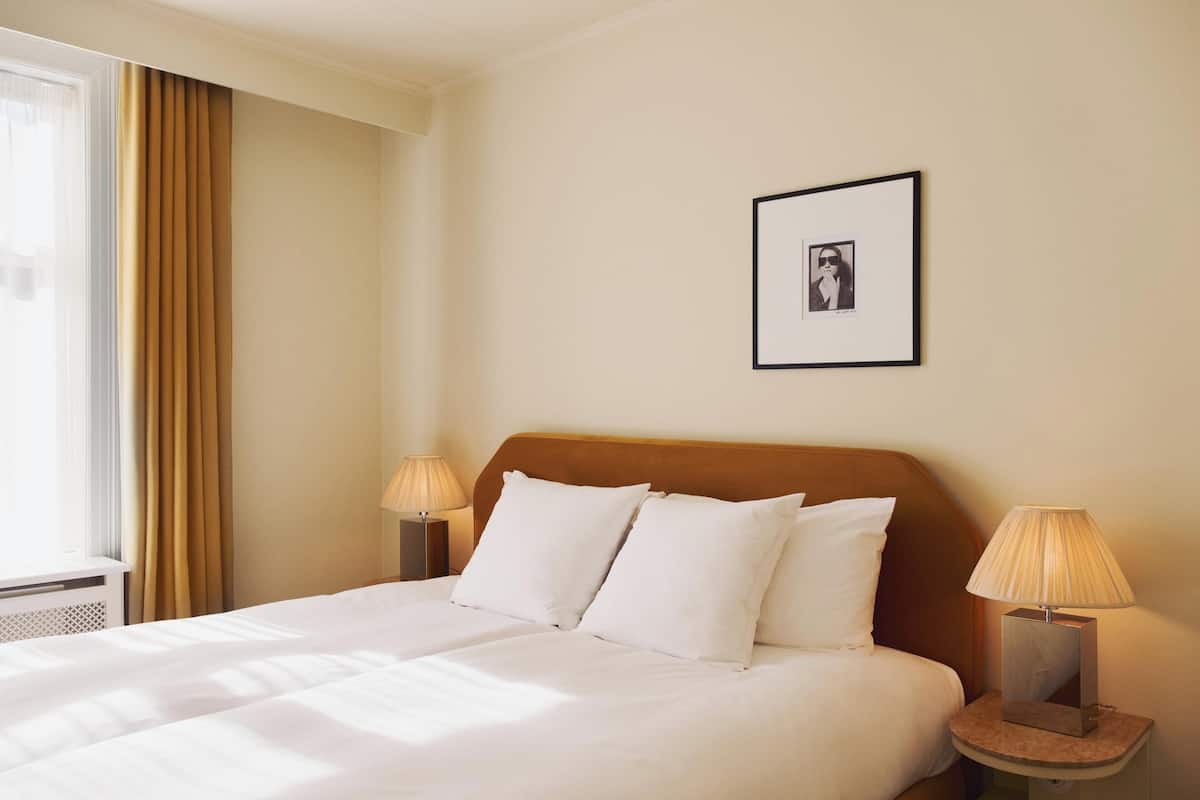 A hotel room with a bed, two bedside lamps, a framed picture on the wall, and a window with curtains.