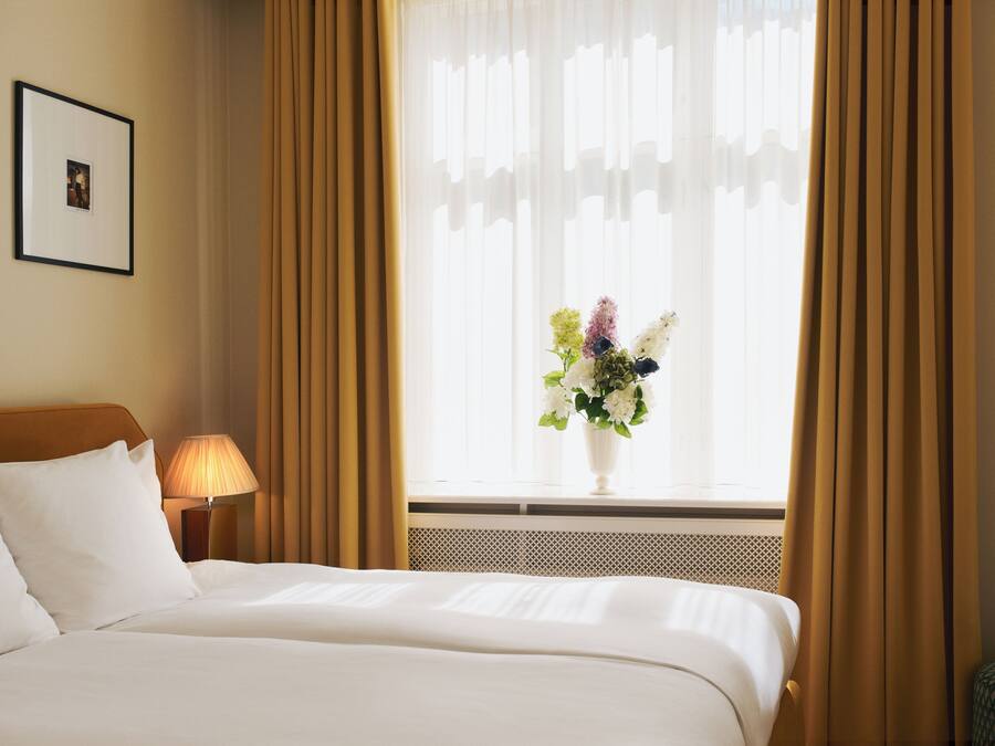 Una habitación de hotel con una cama, un cuadro en la pared, una lámpara y una ventana con cortinas.