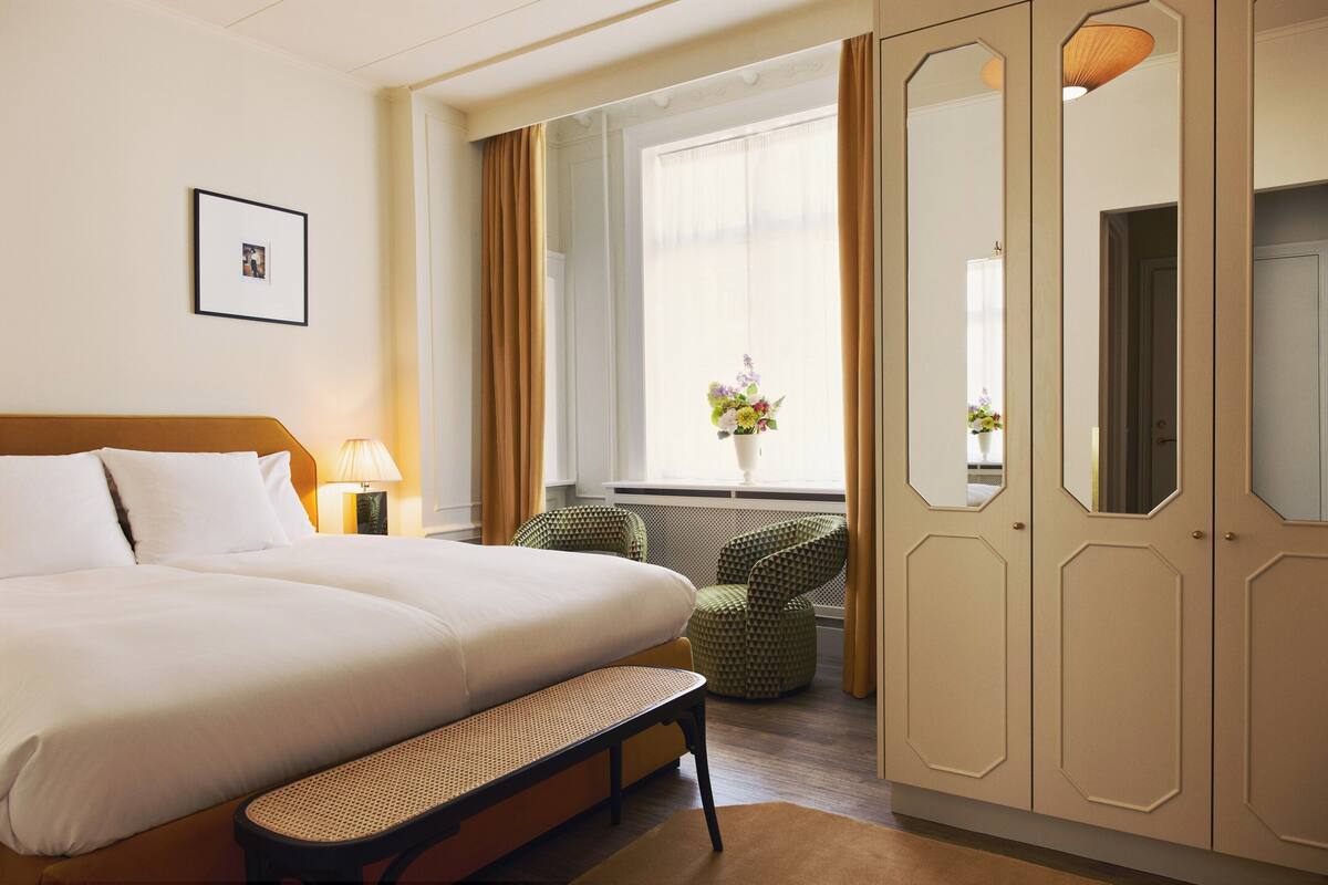 A hotel room with a large bed, a bedside table with a lamp, a bench, a chair, and a wardrobe.