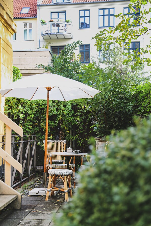 A patio with wicker chairs and a table under a white umbrella, surrounded by greenery and buildings in the background. - Coco Hotel (Copenhagen)