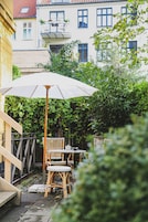 A patio with wicker chairs and a table under a white umbrella, surrounded by greenery and buildings in the background.