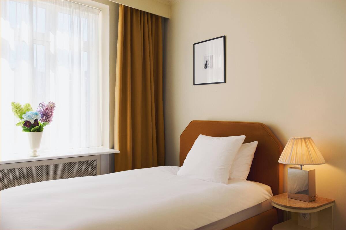 A hotel room with a bed, a bedside table with a lamp, a window with curtains, and a framed picture on the wall.