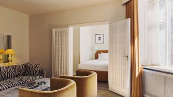 Habitación de hotel con sofá, sillones, cama, ventana con cortinas, lámpara y un cuadro en la pared.