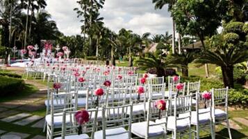 Rows of white chairs with pink flowers, arranged outdoors with a garden and palm trees in the background.