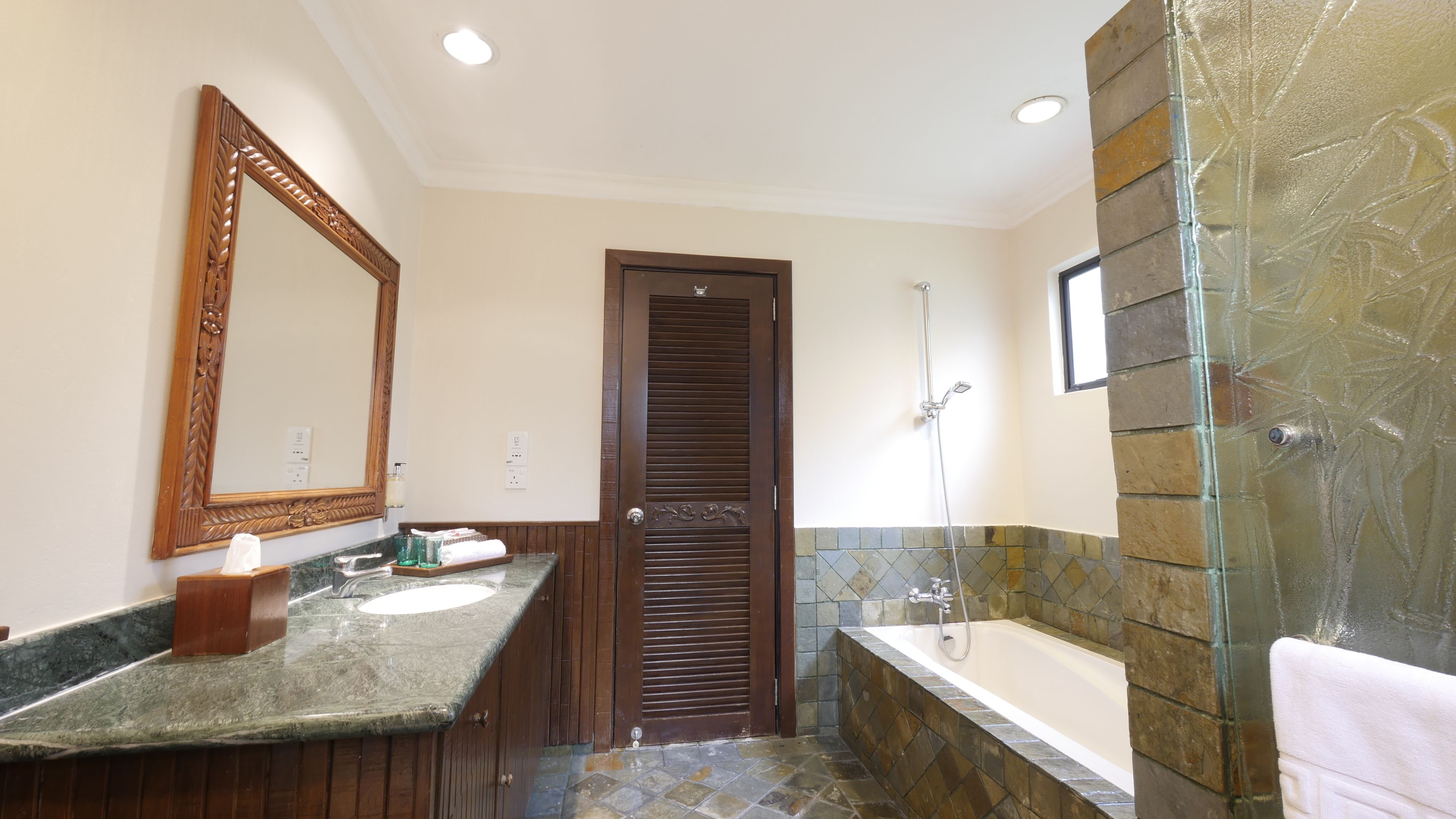 Un baño con puerta de madera, un espejo grande, encimera de mármol, bañera y zona de ducha.