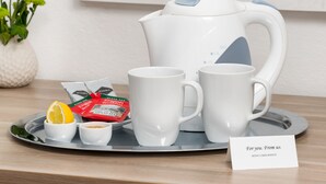 Una bandeja con un hervidor blanco, dos tazas blancas, un cuenco pequeño con una rodaja de limón y un paquete de té. También se ve una tarjeta con el texto 