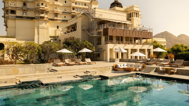 Un área de piscina con sillones reclinables y sombrillas, un edificio grande con varios balcones, y un paisaje montañoso al fondo.
