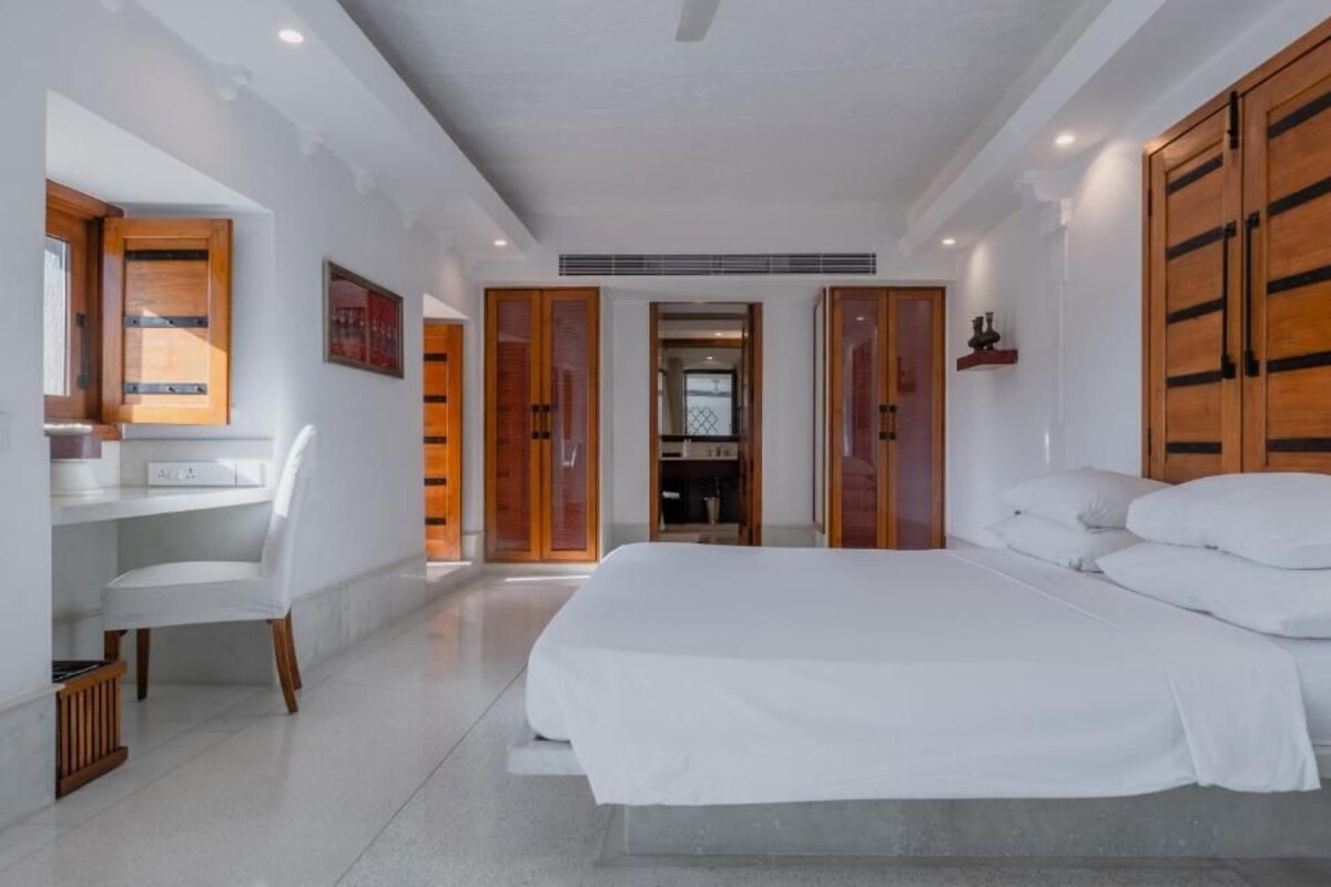 A modern bedroom with a large bed, a desk, and a chair. There are wooden doors and a small shelf.