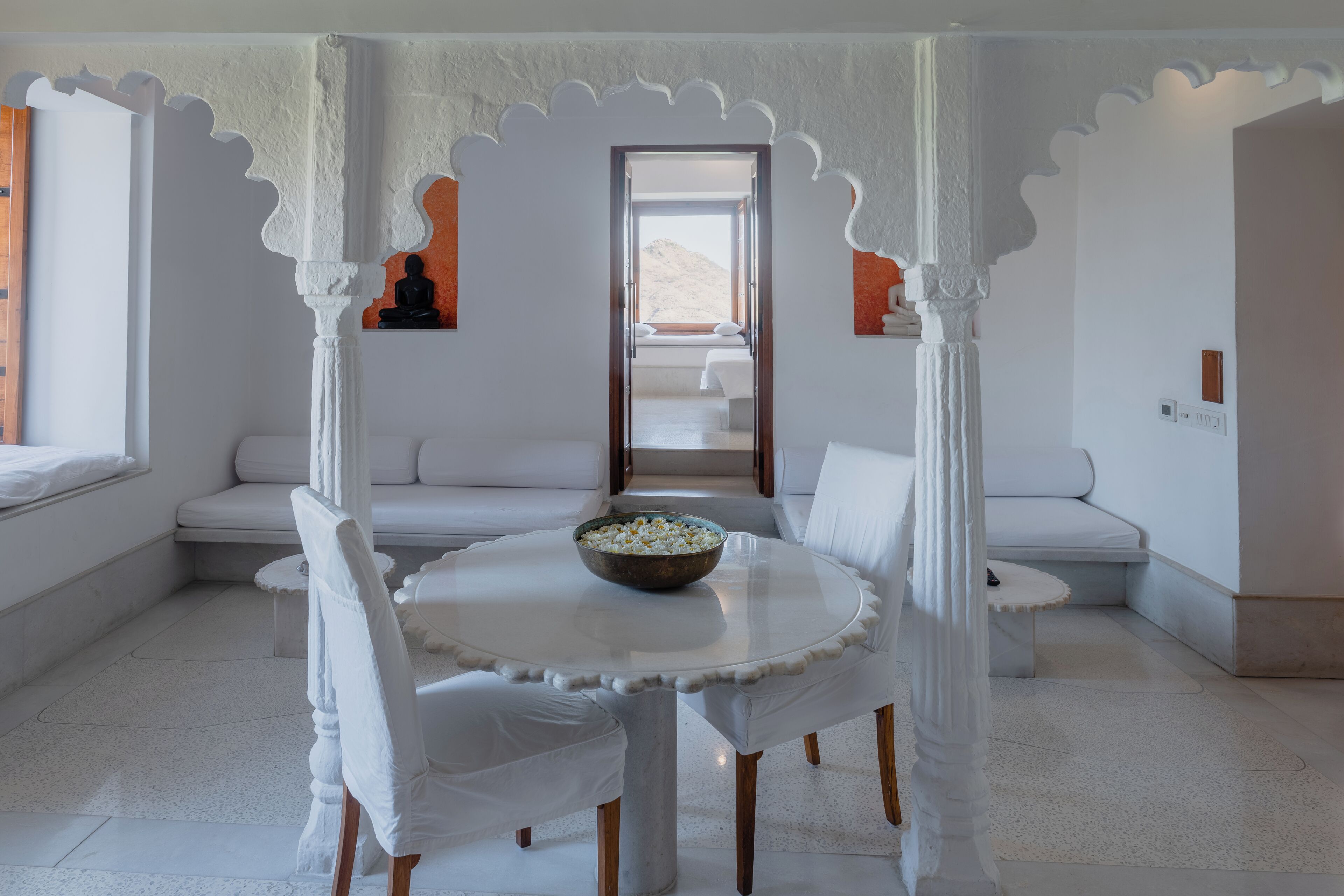 Una habitación con una mesa redonda, sillas blancas, un sofá blanco y vistas a un paisaje montañoso a través de un vano abierto.