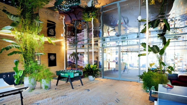 A modern indoor space with a herringbone floor, indoor plants, a foosball table, and a glass wall with reflections. - CPH Hotel (Copenhagen)