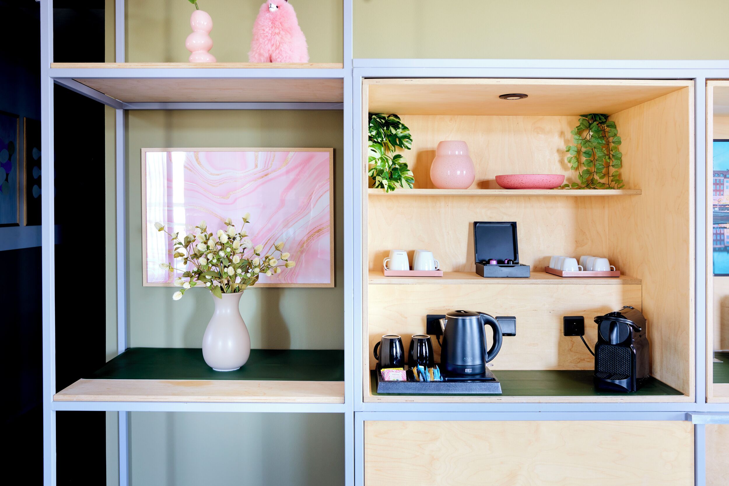 Un espacio habitable compacto con una estación de café, una obra de arte enmarcada y un jarrón rosa con flores.