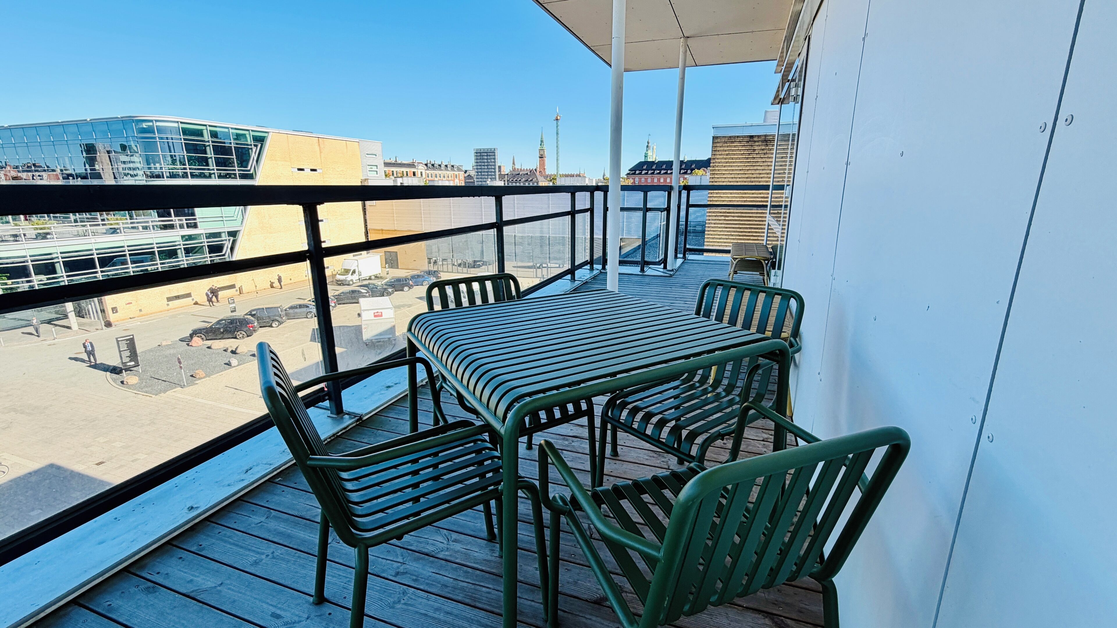 Un balcón con mesa y sillas, con vista a un paisaje urbano de edificios y cielo despejado.