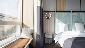 A modern hotel room with a large window, a bed with a headboard, a small bedside table, and a wall-mounted light fixture. - CPH Hotel (Copenhagen)